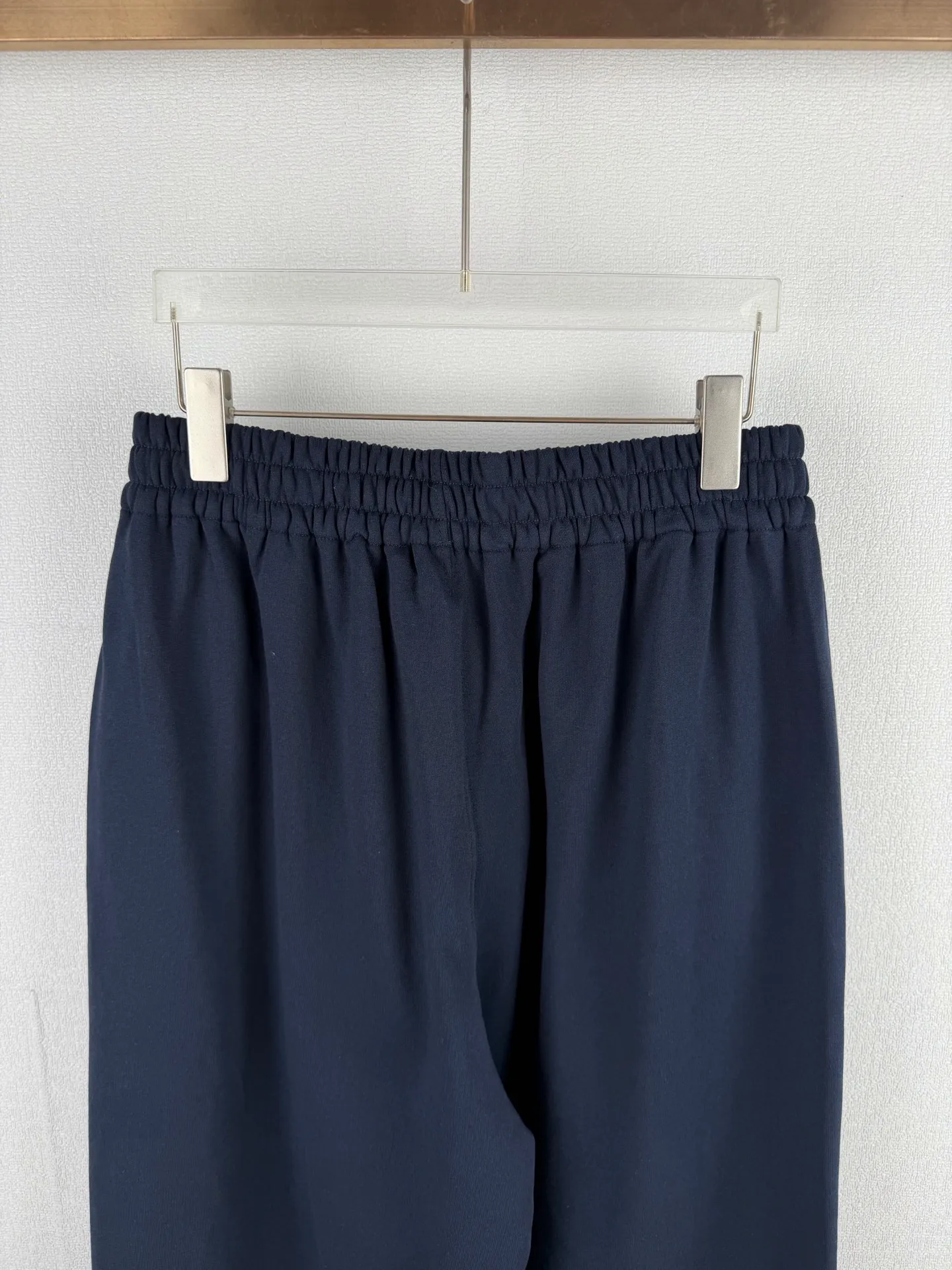 ACNE STUDIOS embroidered logo fleece sweatpants wide-leg trousers. Fabric: 32-count double yarn top + 16-count terry bottom yarn G The veil is made of 32-count fine yarn. The high-count yarn is smooth and delicate. After weaving, it forms a smooth and compact surface. It not only resists pilling and wear-resistant, but also gives the fabric a touch as delicate as skin. The base yarn is made of 16-count roving. The low-count yarn is fluffy and hairy. It naturally forms a rich terry layer during the weaving process, providing a sufficient source of fiber for subsequent sanding. This combination of fine surfaces and thick soles strikes a perfect balance between warmth and skin-friendliness. Mechanical sanding is the soul process of this fabric. By rubbing the bottom surface of the roller wrapped with emery leather at high speed, the fiber ends of the 16-count roving are evenly pulled out and cut off, forming a dense and uniform short pile layer. The smoothness and neatness of needle embroidery and the soft velvet feel of brushed fabrics form a material dialogue between hardness and softness. The embroidery area is smooth and crisp, with sharp lines, like delicate inscriptions carved on a rich suede canvas; while the surrounding brushed fabric presents a soft matte natural texture. The spacing between stitches is consistent and the strength is balanced to ensure that each stitch is placed accurately. This rigorous stitching structure makes the edges of the logo present a sharp and clear outline, and the strokes of the text are full and smooth. Even the curved turning points of the floral fonts can achieve a smooth transition through the connection of tight stitches.